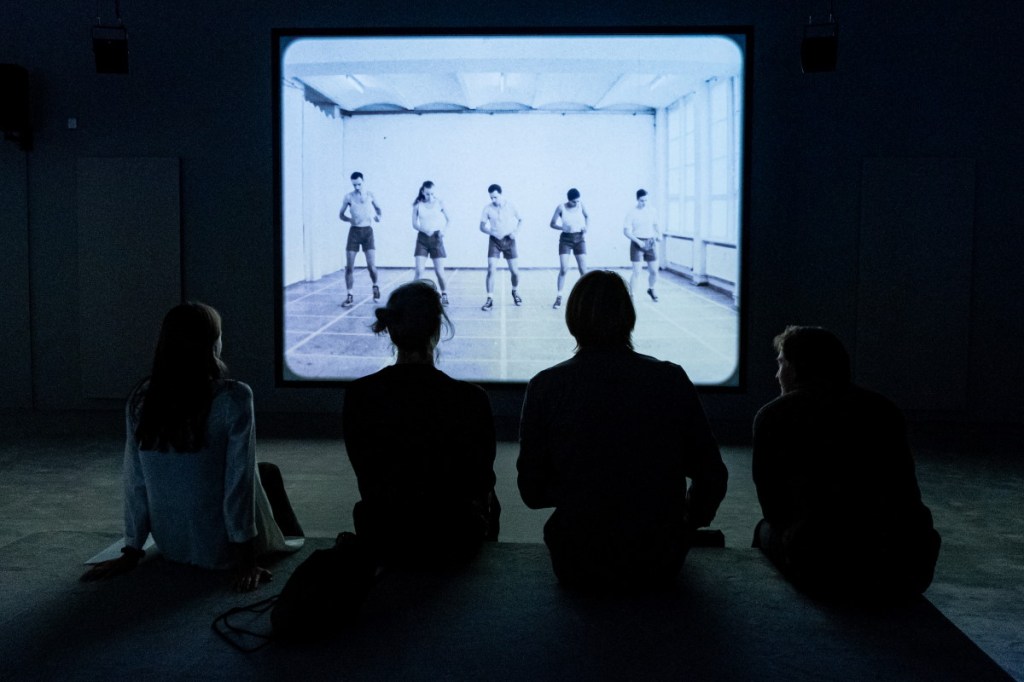 neli inimest istub Kumu suures saalis mustvalge videoga ekraani ees